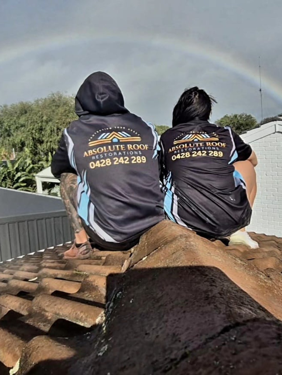 Two of the crew sitting up on the ridge in Absolute Roof Restorations shirts, a rainbow behind them